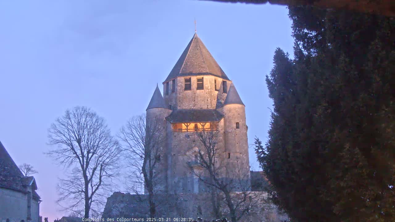A stone tower, illuminated from within, stands under a twilight sky, surrounded by bare trees and evergreen shrubs.