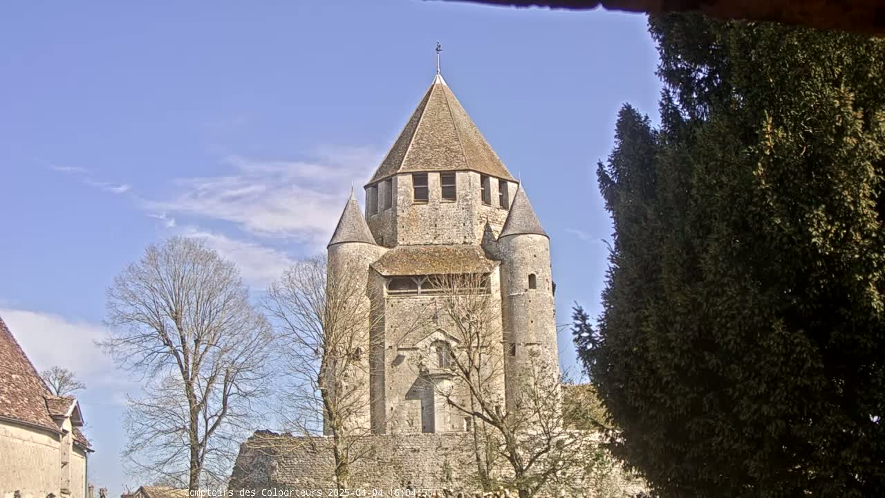 A tall, stone tower with a conical roof and smaller flanking towers stands under a mostly clear, blue sky, partially obscured by bare trees and an evergreen tree.