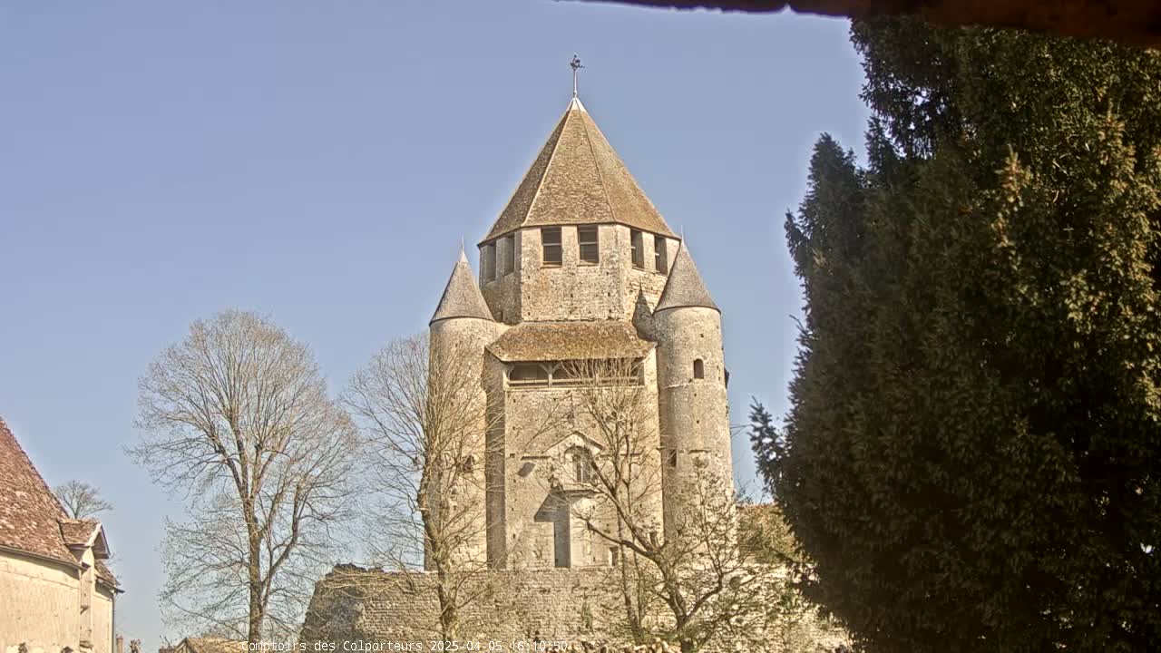 A large, stone tower with a conical roof stands among bare trees under a clear blue sky.