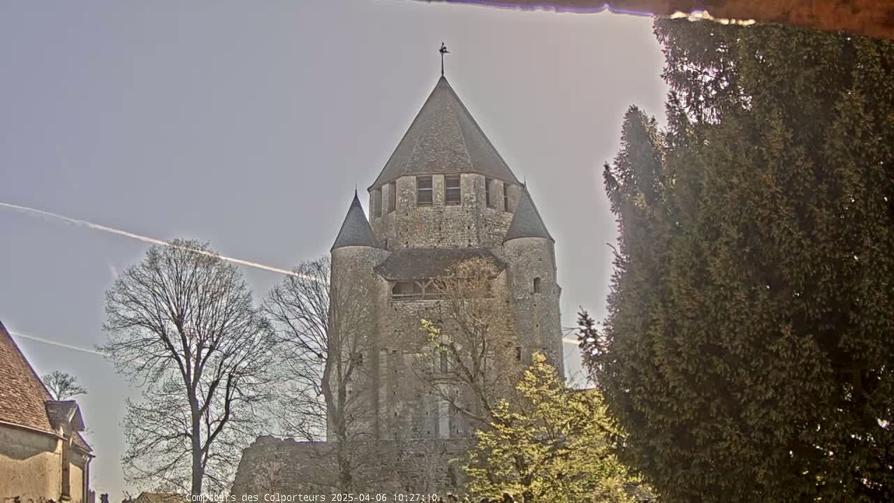 A tall, stone tower with a conical roof stands amidst bare and budding trees under a pale blue, mostly clear sky with a few contrails.