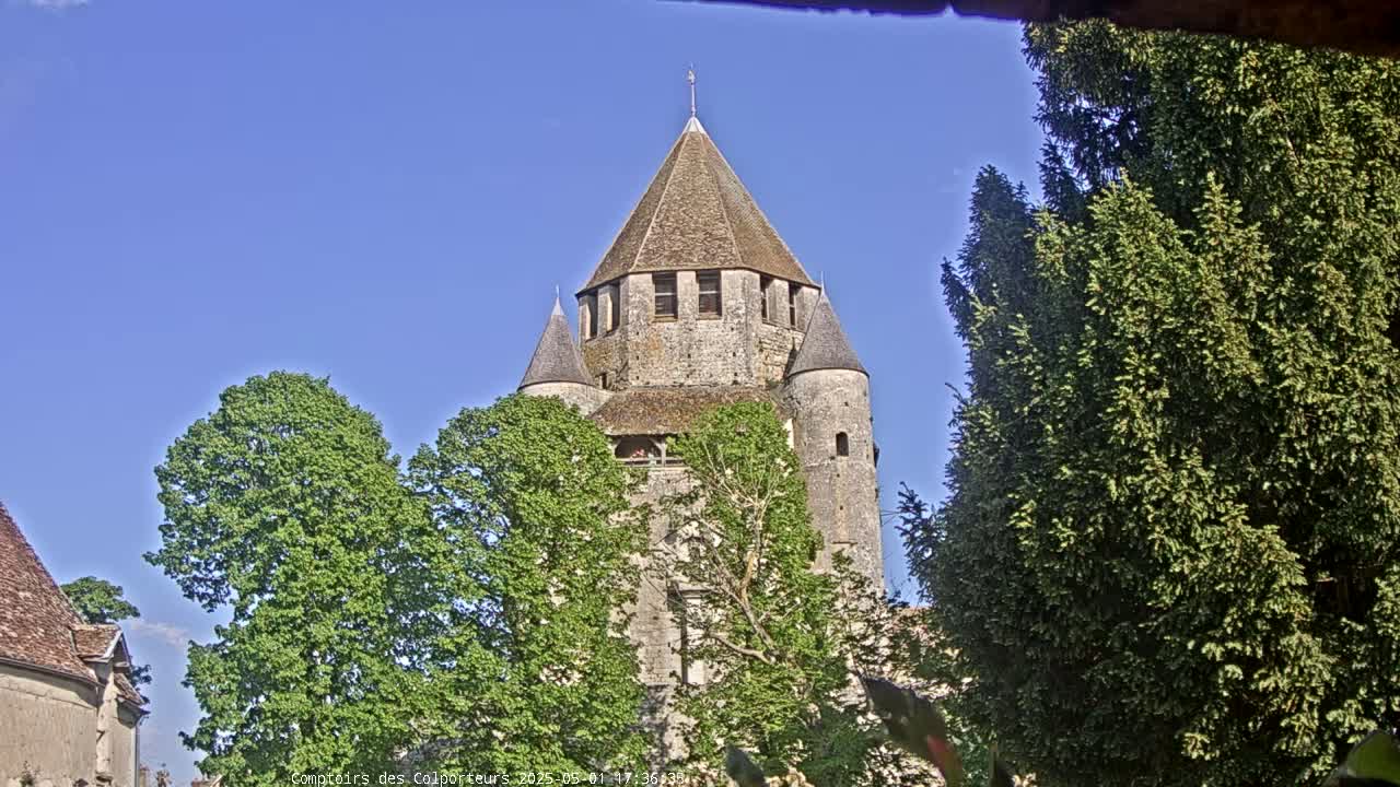 A stone tower with a conical roof is partially obscured by lush green trees under a clear blue sky.