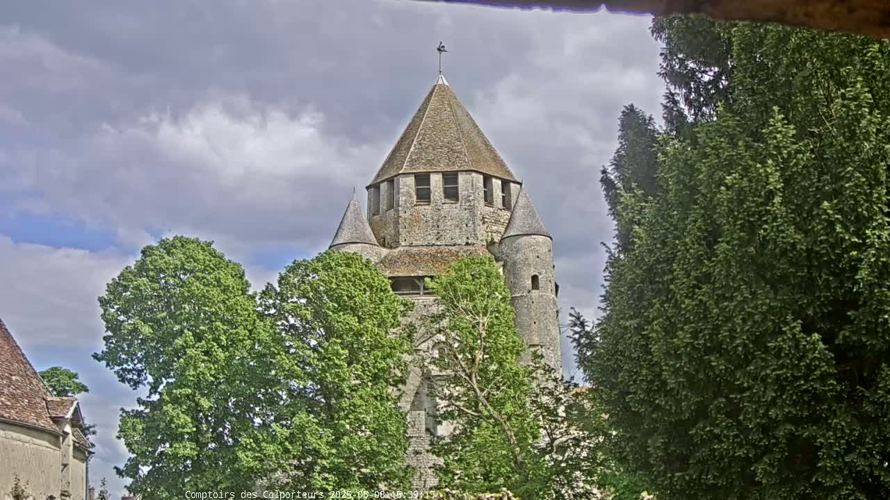 A stone tower with a conical roof, partially obscured by trees, stands under a partly cloudy sky.