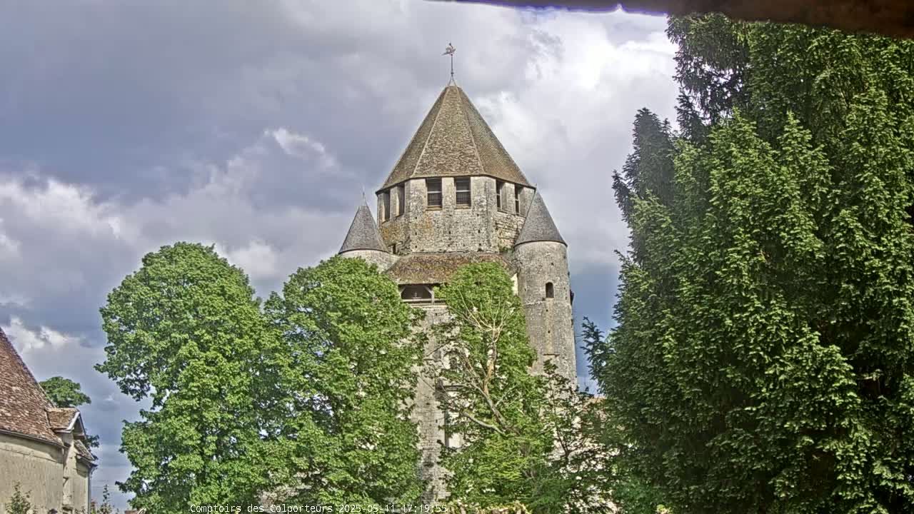 A stone tower with a conical roof is partially obscured by green trees under a partly cloudy sky.