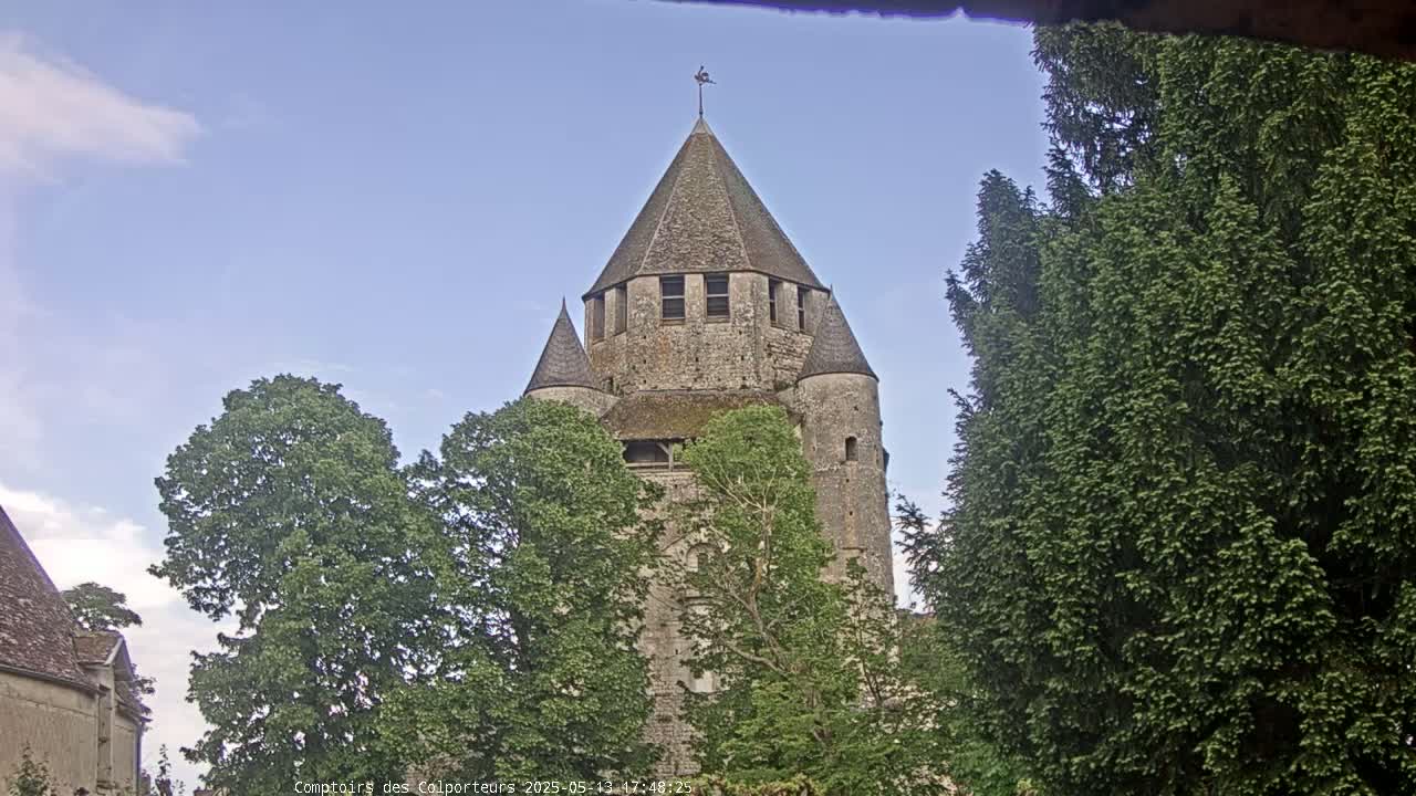 A stone tower, partially obscured by leafy green trees, stands under a mostly clear blue sky.