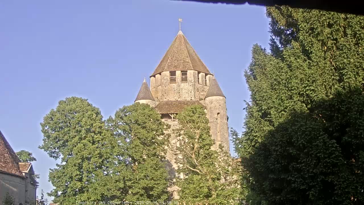 A stone tower with a conical roof is partially obscured by leafy green trees under a clear blue sky.