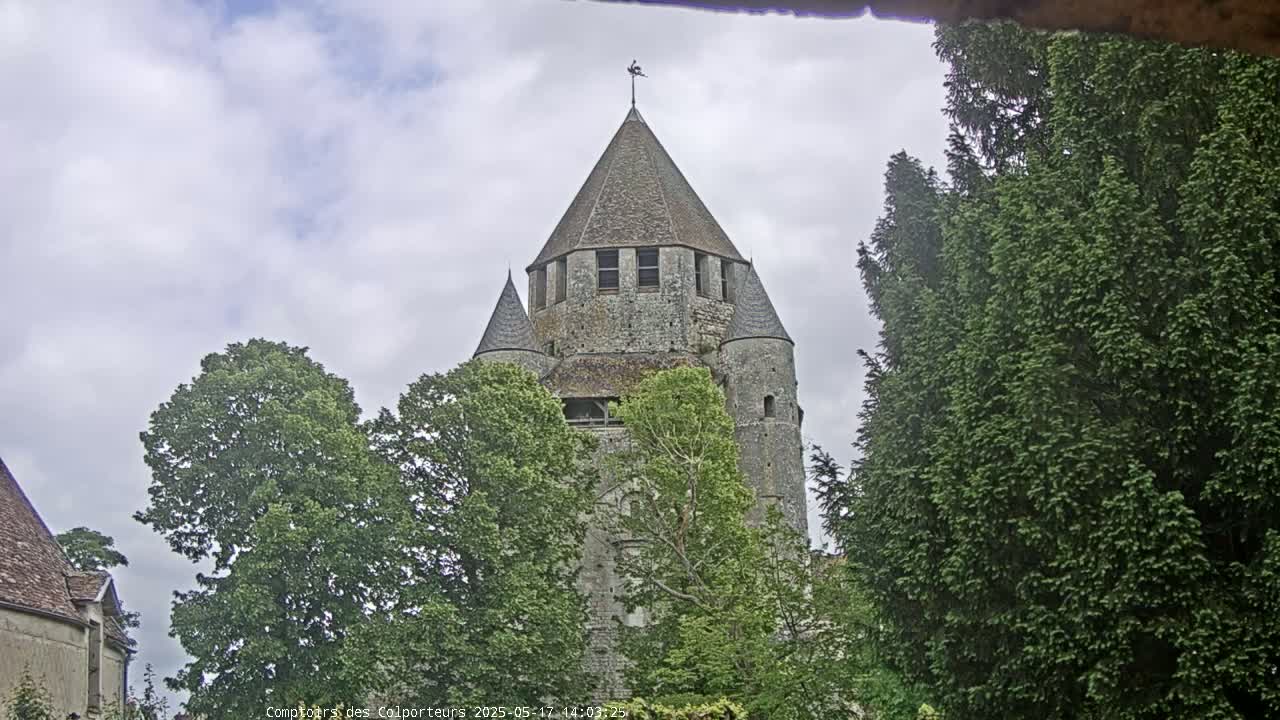 A stone tower with a conical roof is partially obscured by lush green trees under a cloudy sky.