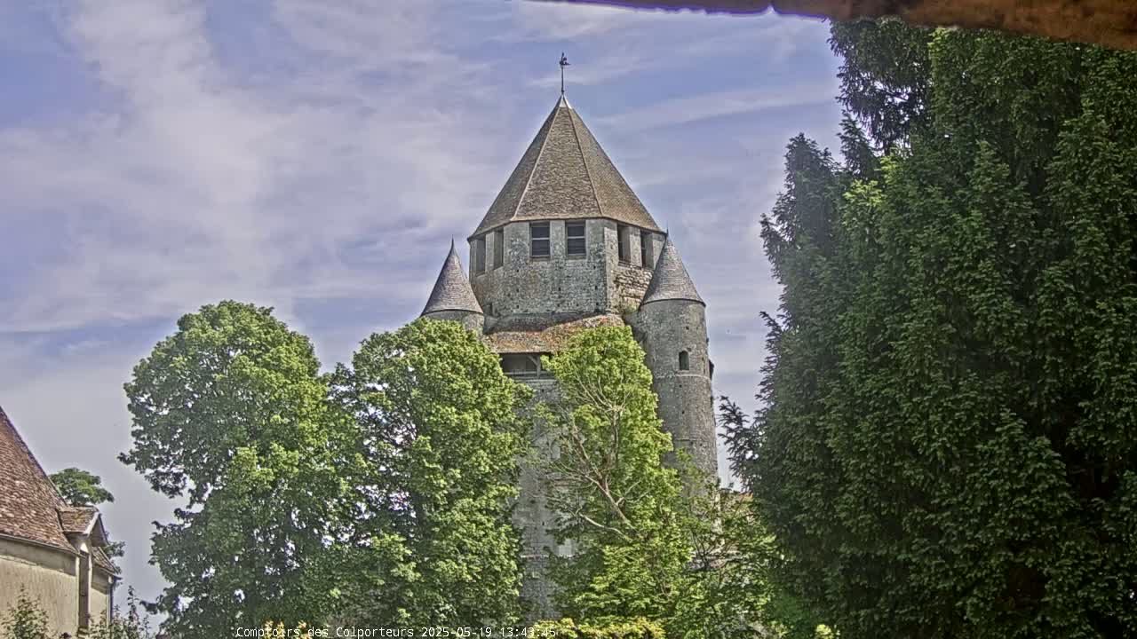 A stone tower with conical roofs, partially obscured by green trees, stands under a partly cloudy sky.