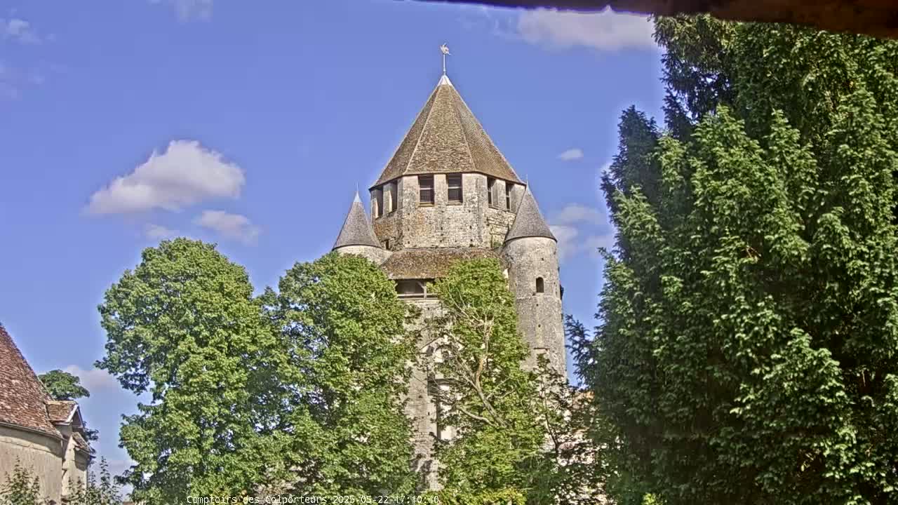 A stone tower with a conical roof, flanked by smaller turrets, is partially obscured by lush green trees under a mostly sunny sky with a few scattered clouds.