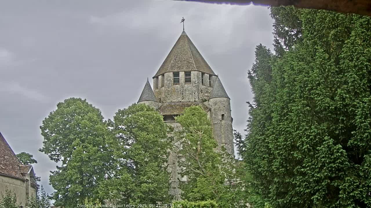 A stone tower with a conical roof is partially obscured by lush green trees under a cloudy sky.