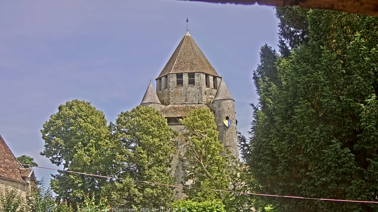 A stone tower with a conical roof is partially obscured by lush green trees under a clear blue sky.