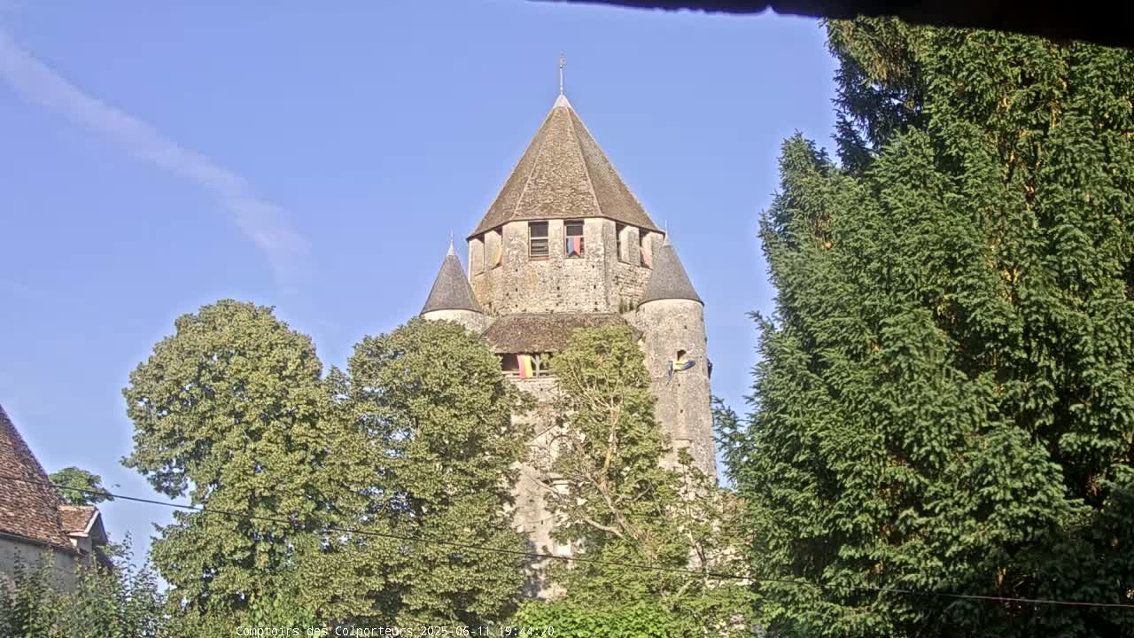 A stone tower with a conical roof is seen partially obscured by trees under a clear blue sky.
