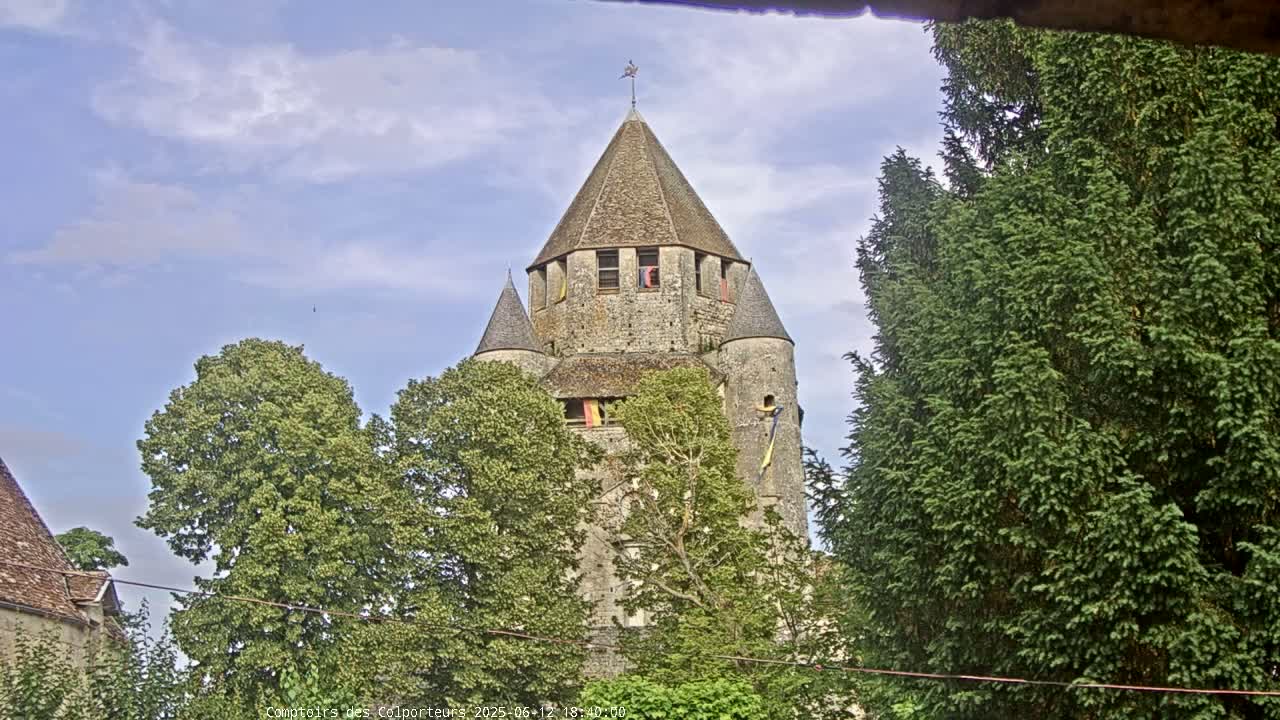 A stone tower with a conical roof is partially obscured by lush green trees under a partly cloudy sky.