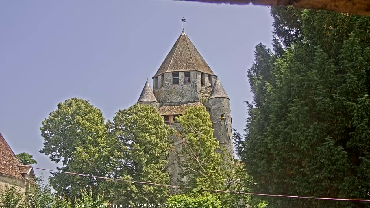 A stone tower with a conical roof is partially obscured by lush green trees under a clear blue sky.