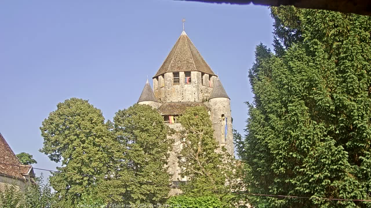 A stone tower with a conical roof is partially obscured by lush green trees under a clear blue sky.