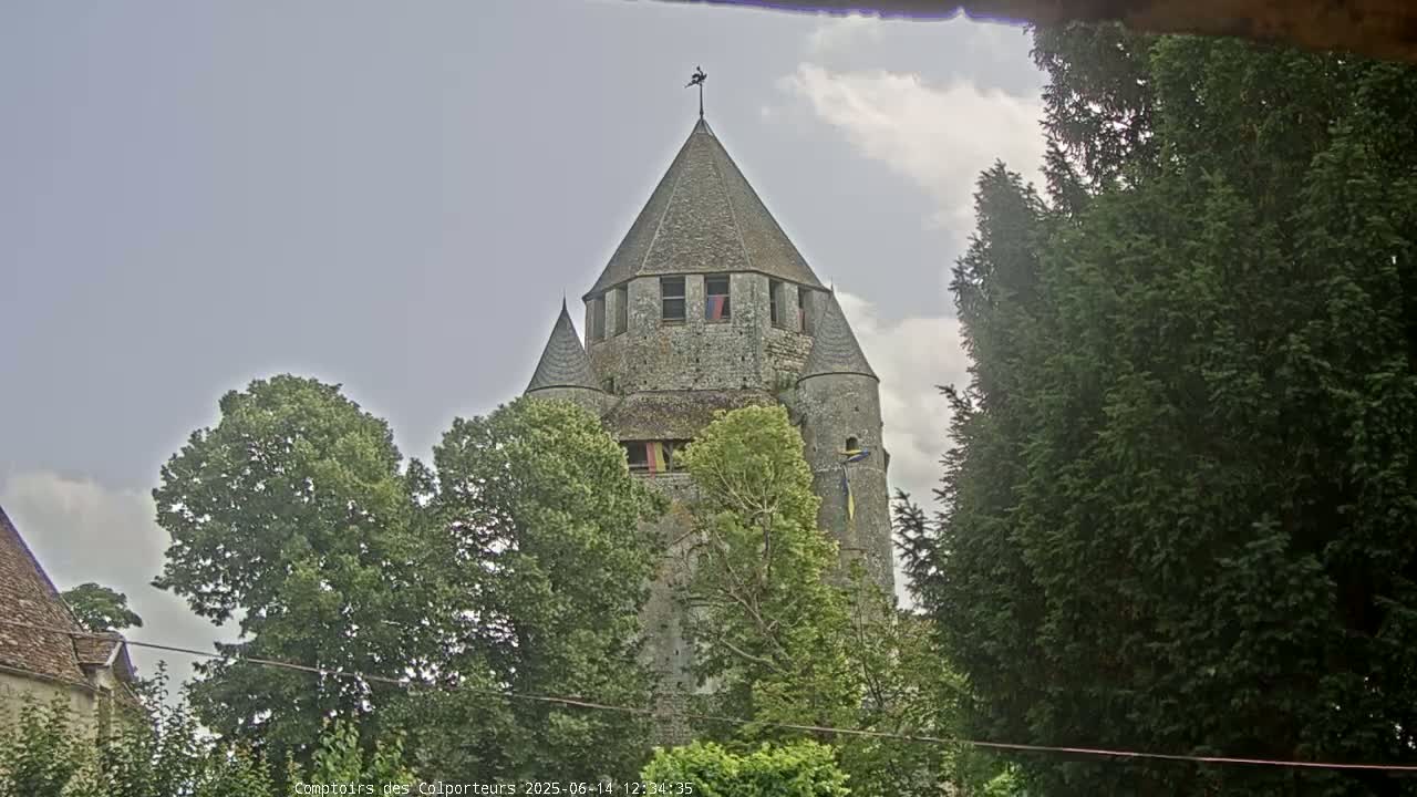 A stone tower with a conical roof is partially obscured by lush green trees under a cloudy sky.