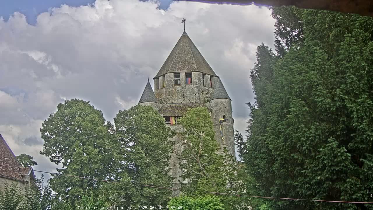 A stone tower with a conical roof stands amidst green trees under a partly cloudy sky.