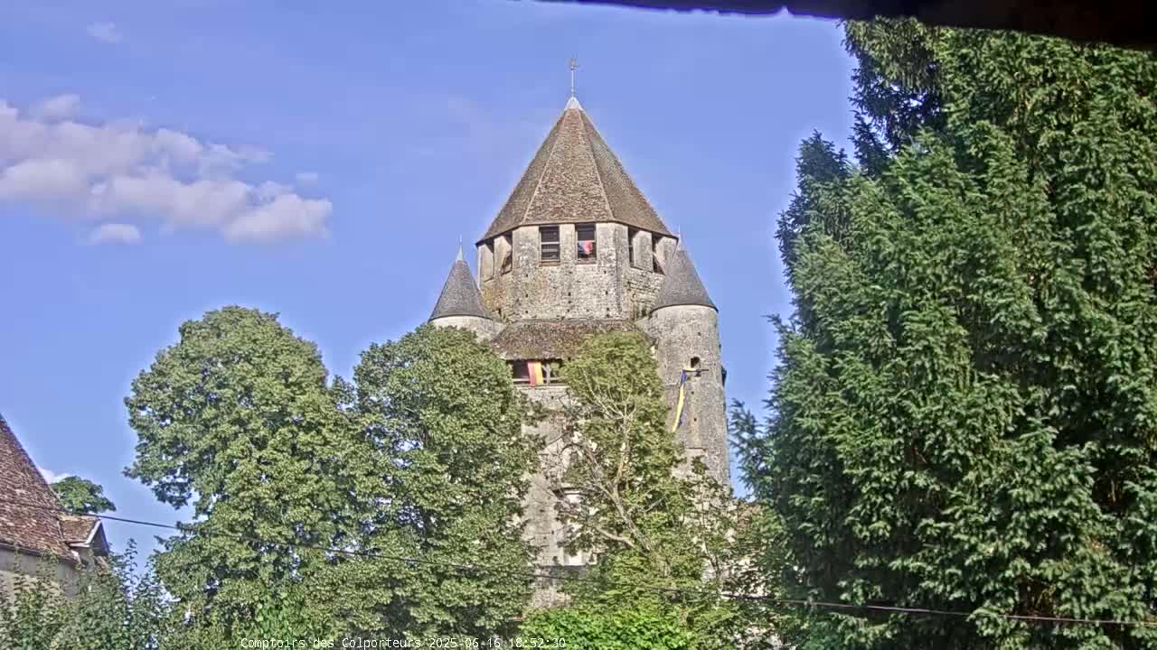 A stone tower with a conical roof is partially obscured by lush green trees under a mostly clear blue sky.