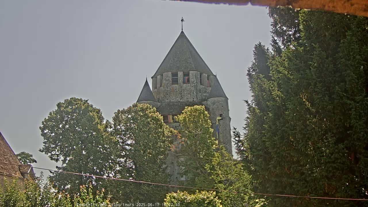 A stone tower with a conical roof is partially visible through a screen of green trees under a clear, light-grey sky.