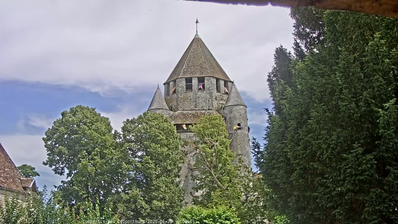 A stone tower with a conical roof is partially obscured by leafy green trees under a partly cloudy sky.