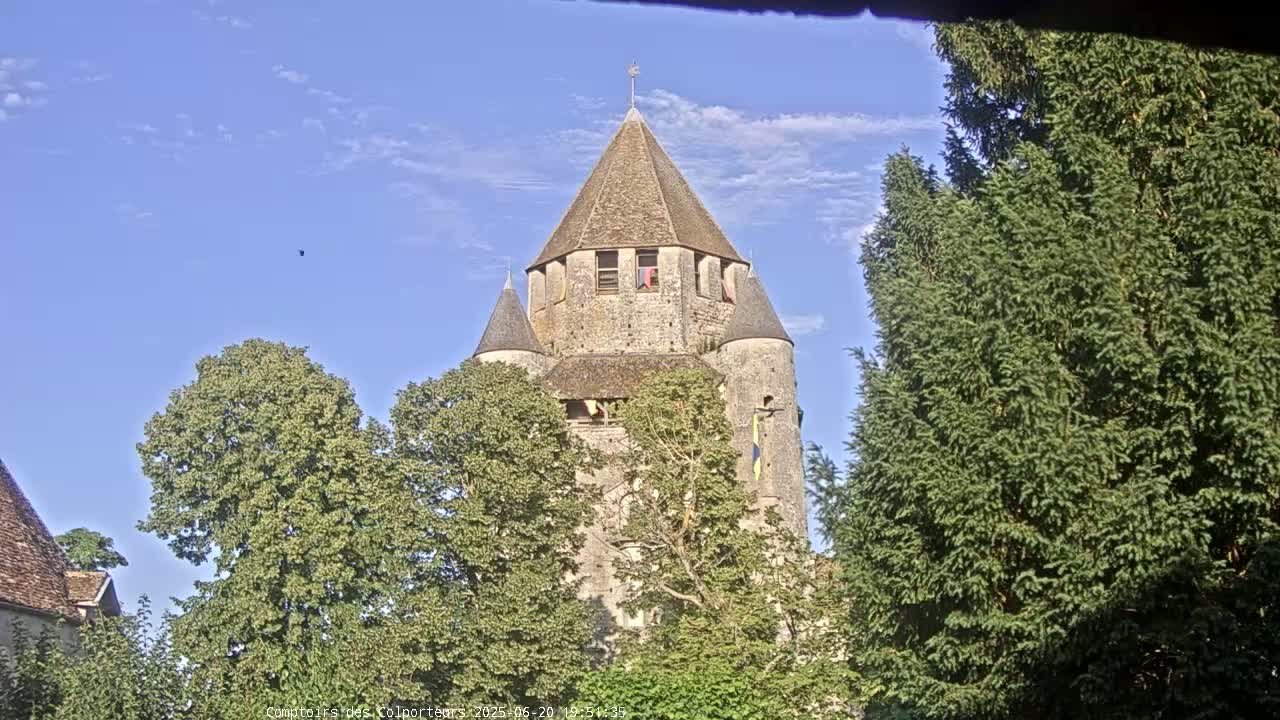 A stone tower with a conical roof is partially obscured by leafy green trees under a mostly clear blue sky.