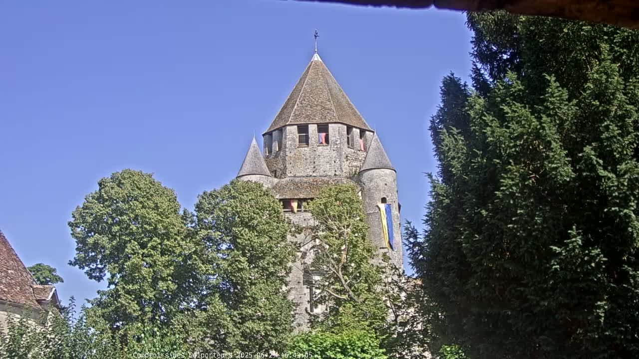 A stone tower with a conical roof is partially obscured by leafy green trees under a clear blue sky.
