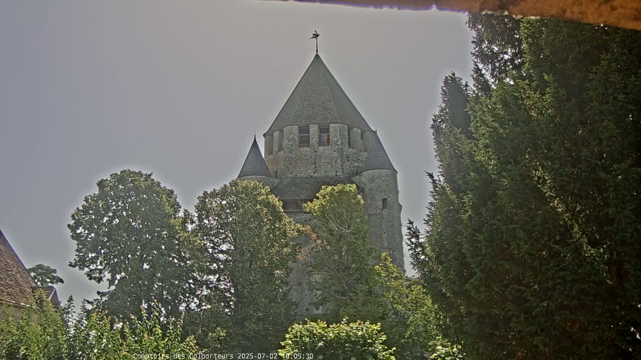 A stone tower with a conical roof rises above leafy green trees under a bright, overcast sky.