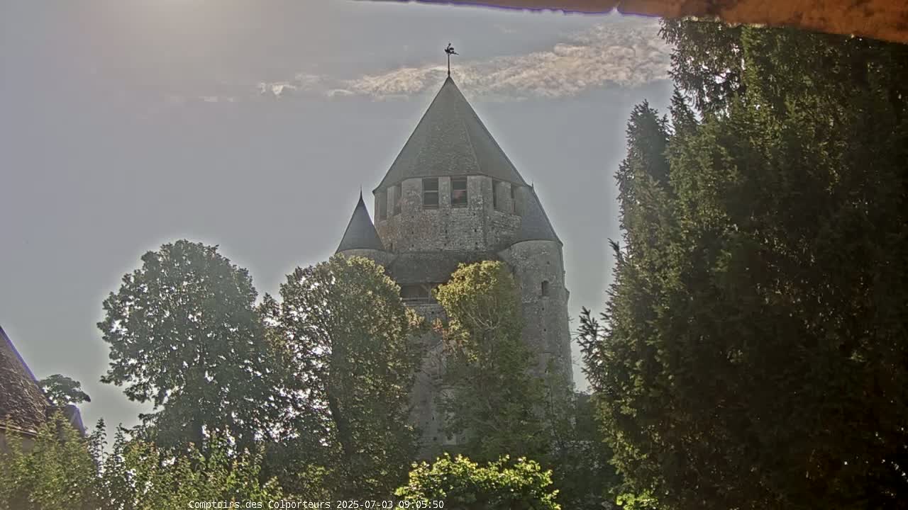 A tall, stone tower with a pointed roof is partially obscured by green trees under a mostly cloudy sky.