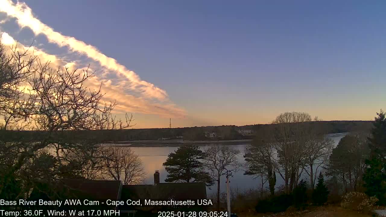 A calm river reflects the sunset under a partly cloudy sky with a distinct band of orange-hued clouds, and bare trees line the riverbanks.