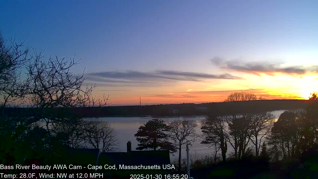 A colorful sunset over a calm river, viewed from a vantage point among bare trees in a cool, windy climate.