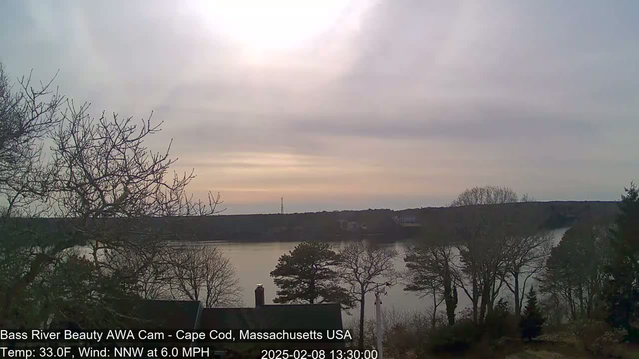 A calm river is seen at sunset under a mostly cloudy sky, surrounded by bare, winter trees.