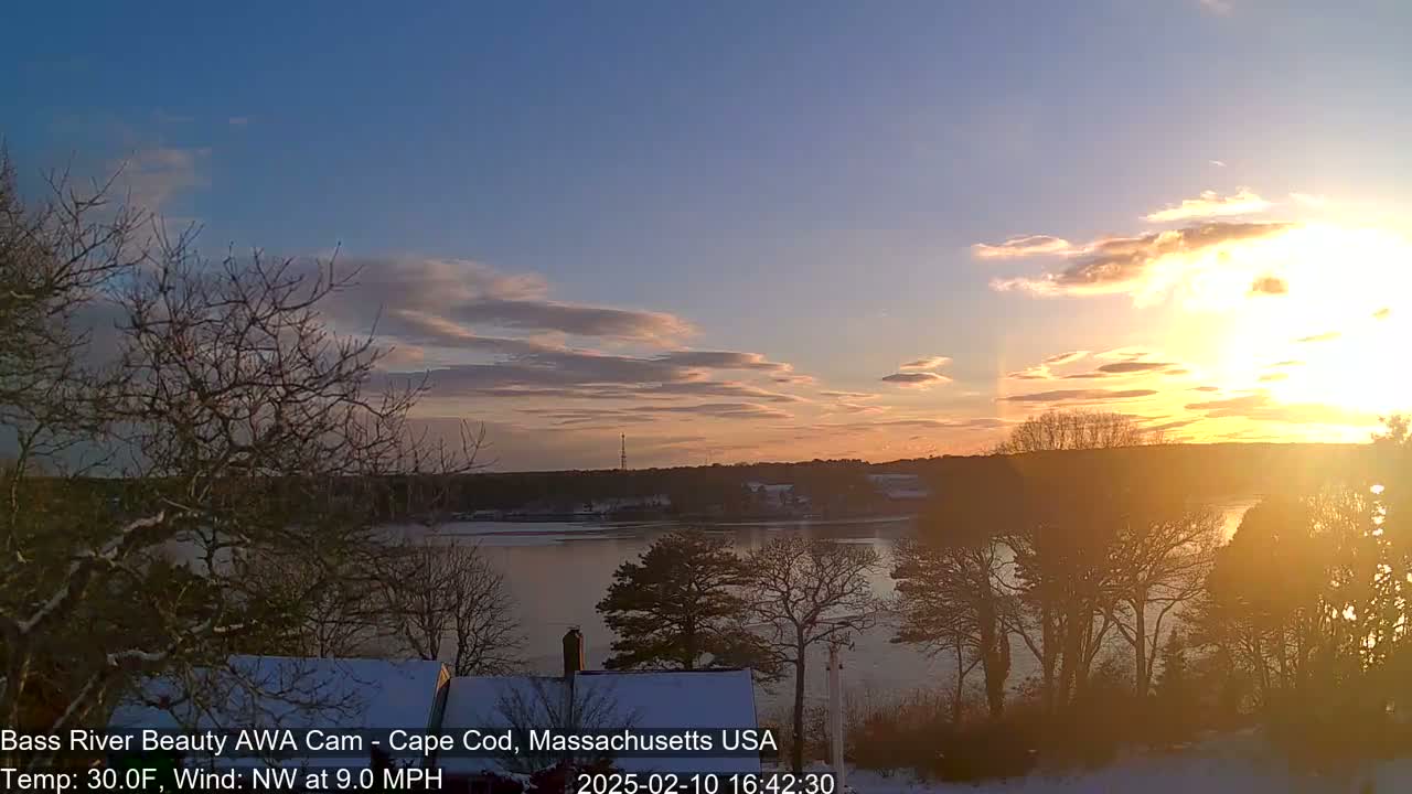 A snow-covered landscape with leafless trees overlooks a partially frozen river at sunset under a partly cloudy sky.
