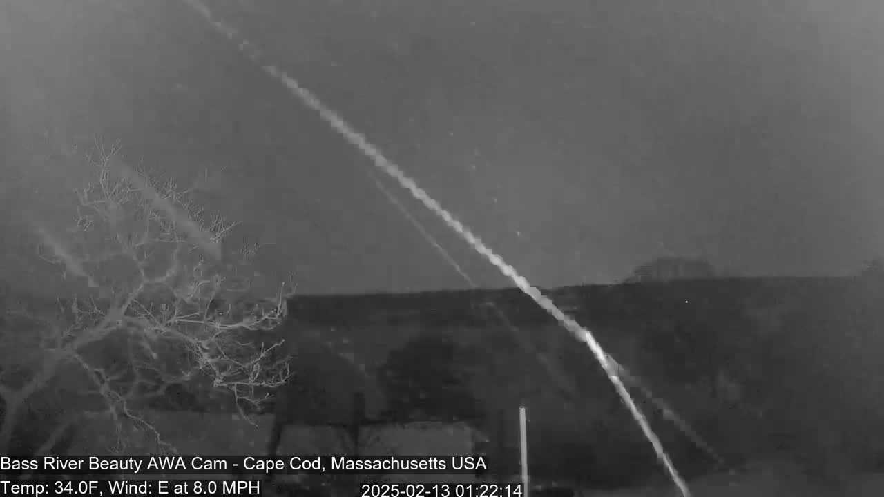 A nighttime, grayscale image shows a barren tree in the foreground with two bright streaks across a dark sky, under 34°F weather conditions with an 8 mph east wind.