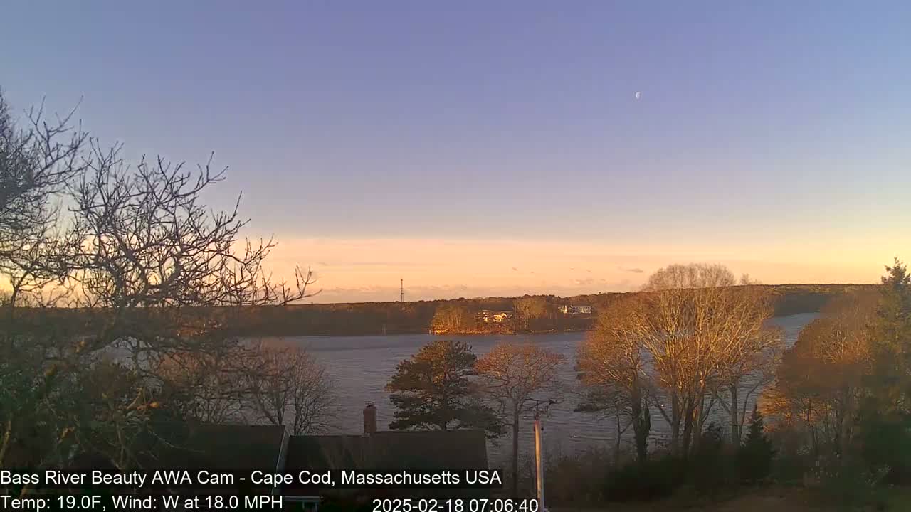 A calm river at sunset under a pale blue sky with a crescent moon, is viewed from a vantage point among leafless trees.