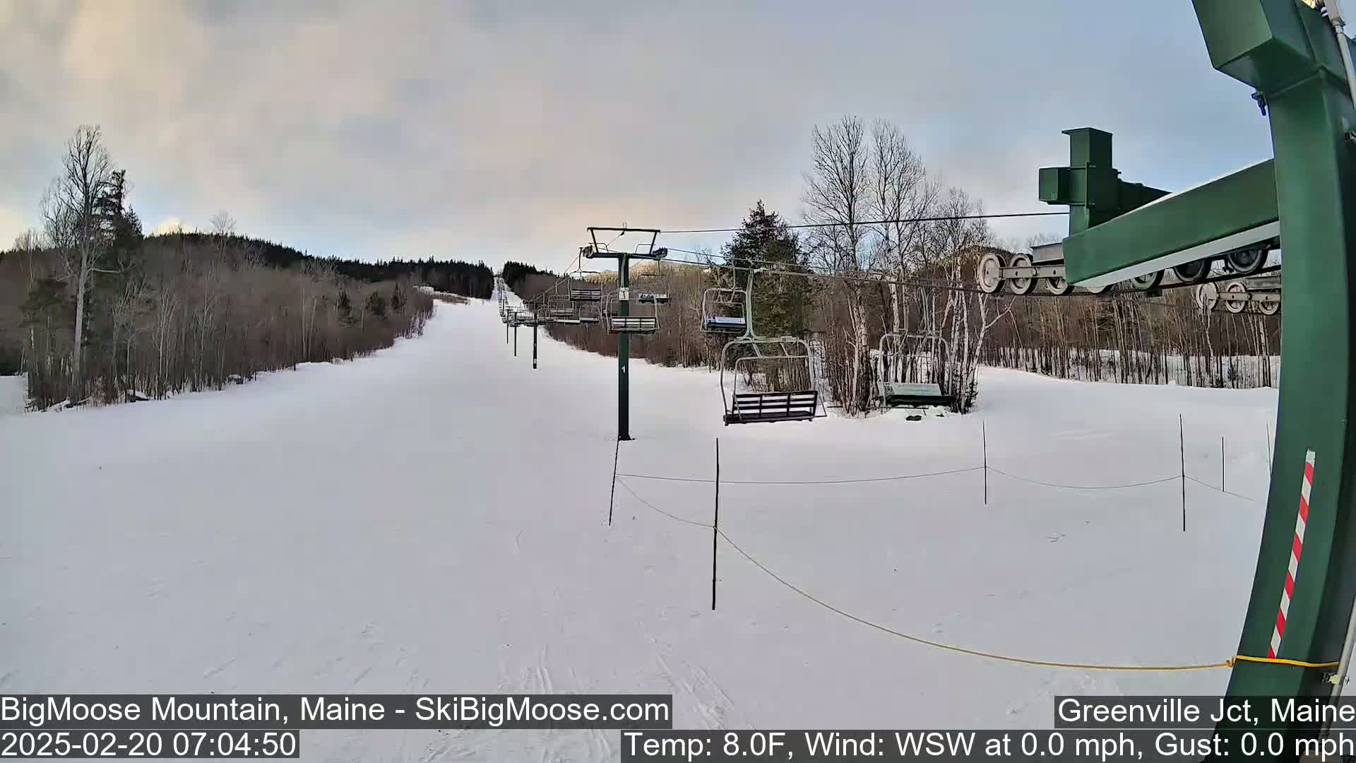 A snow-covered ski slope with a chairlift and sparsely wooded areas under a mostly cloudy sky.