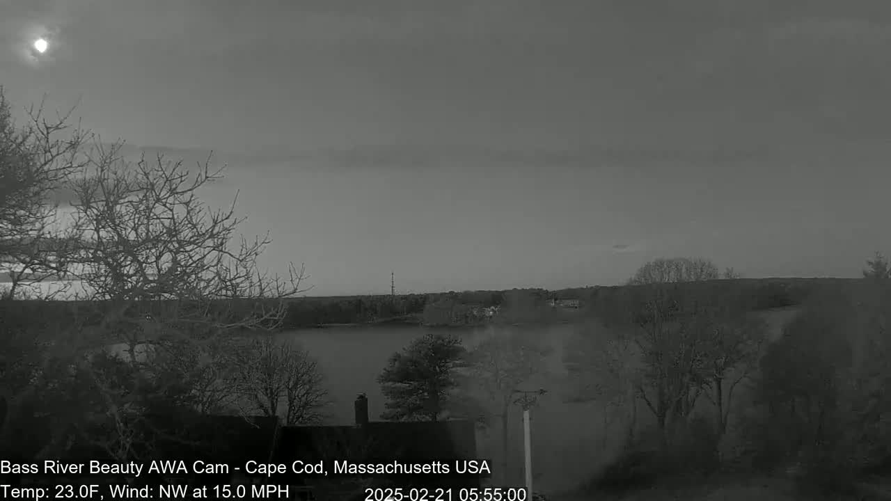 A grayscale image shows a body of water at dusk or dawn under a mostly clear sky with a bright celestial body visible, bare winter trees, and a few buildings in the distance, with a northwest wind at 15 mph and a temperature of 23 degrees Fahrenheit.