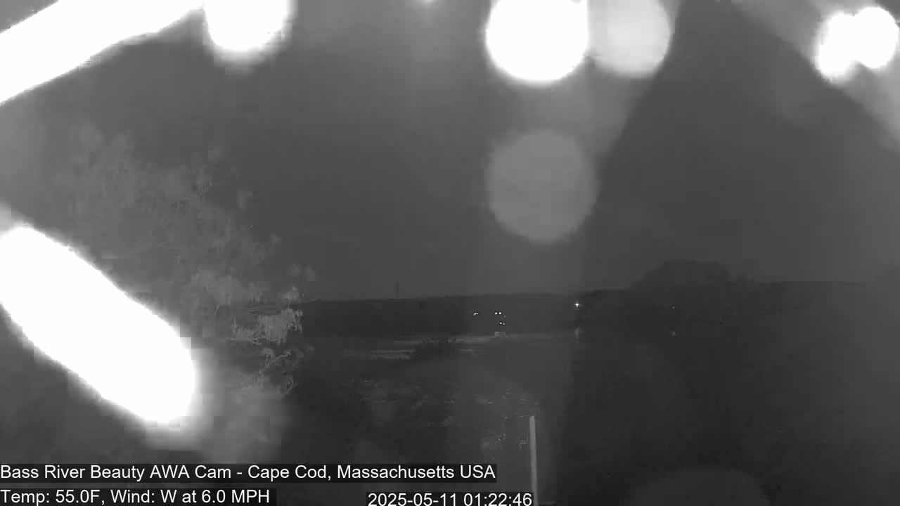 A nighttime, low-light outdoor scene shows blurry lights and a dark landscape under a slightly hazy sky, with a 6.0 mph westerly wind.