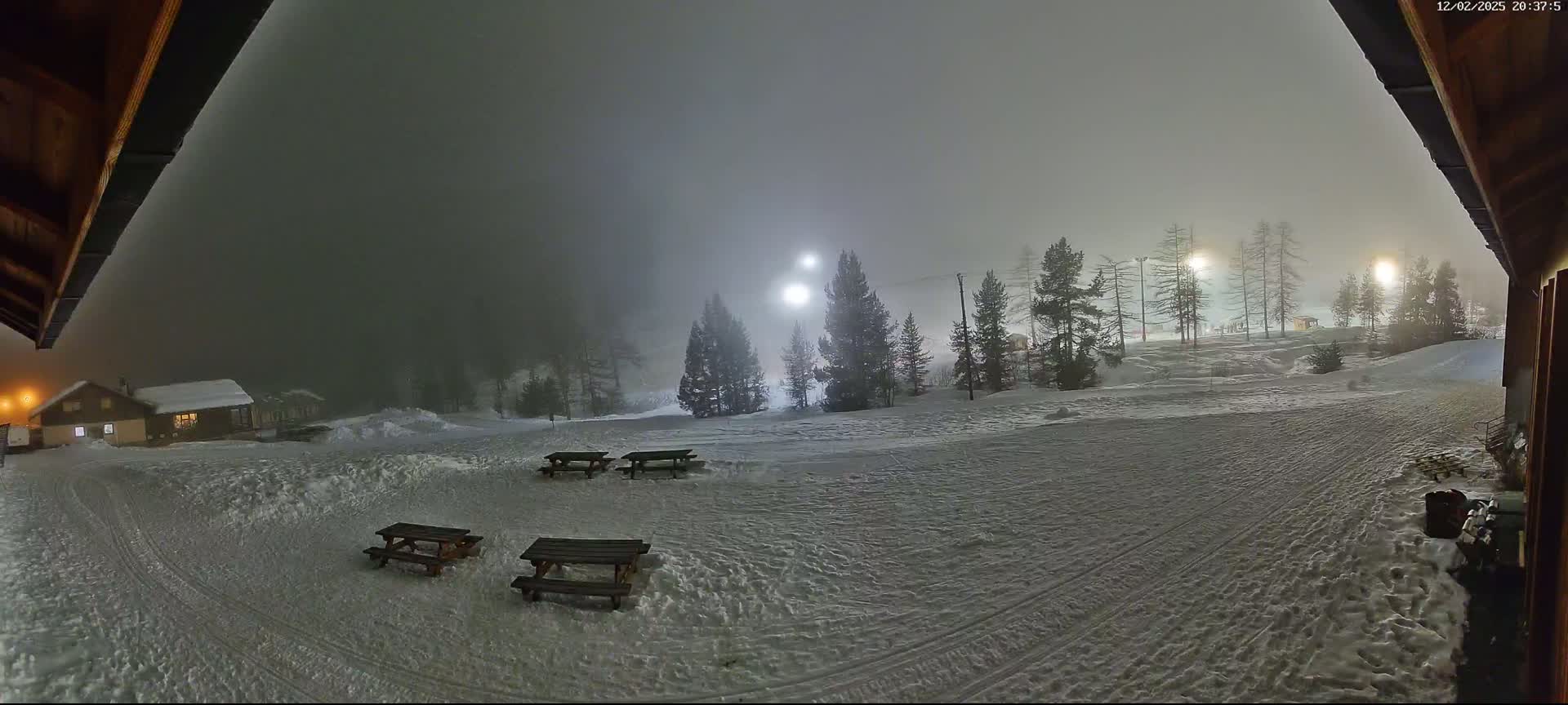 A snowy, foggy night scene shows several picnic tables in a snow-covered area near a building, with illuminated trees and a ski slope in the background.