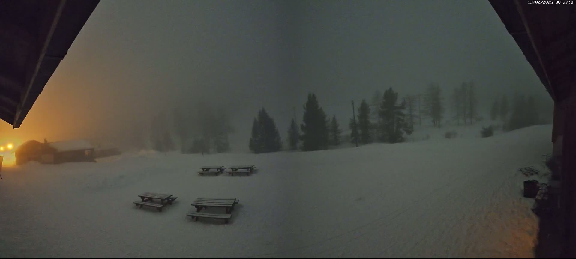 A snow-covered landscape is visible through a doorway at night, with several picnic tables and some distant trees obscured by heavy fog or snow.