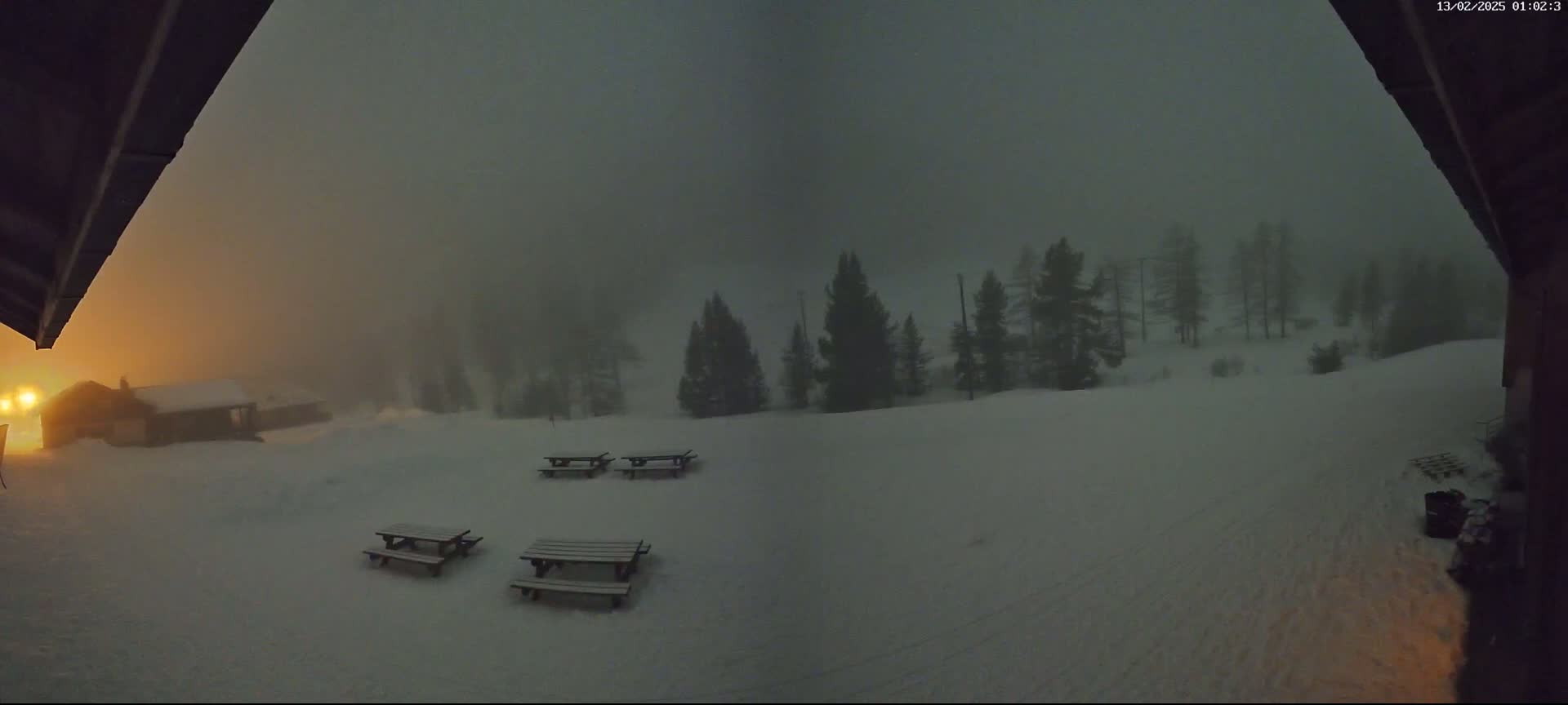 A snowy, foggy scene at night shows several picnic tables in a clearing, a small building in the distance, and dark evergreens on a hill under a dimly lit sky.