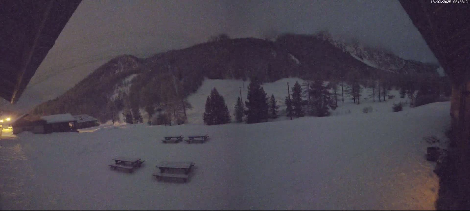 A snow-covered landscape at night, featuring a few picnic tables and a small building in the foreground, with a dark, snow-covered mountain in the background under a cloudy sky.