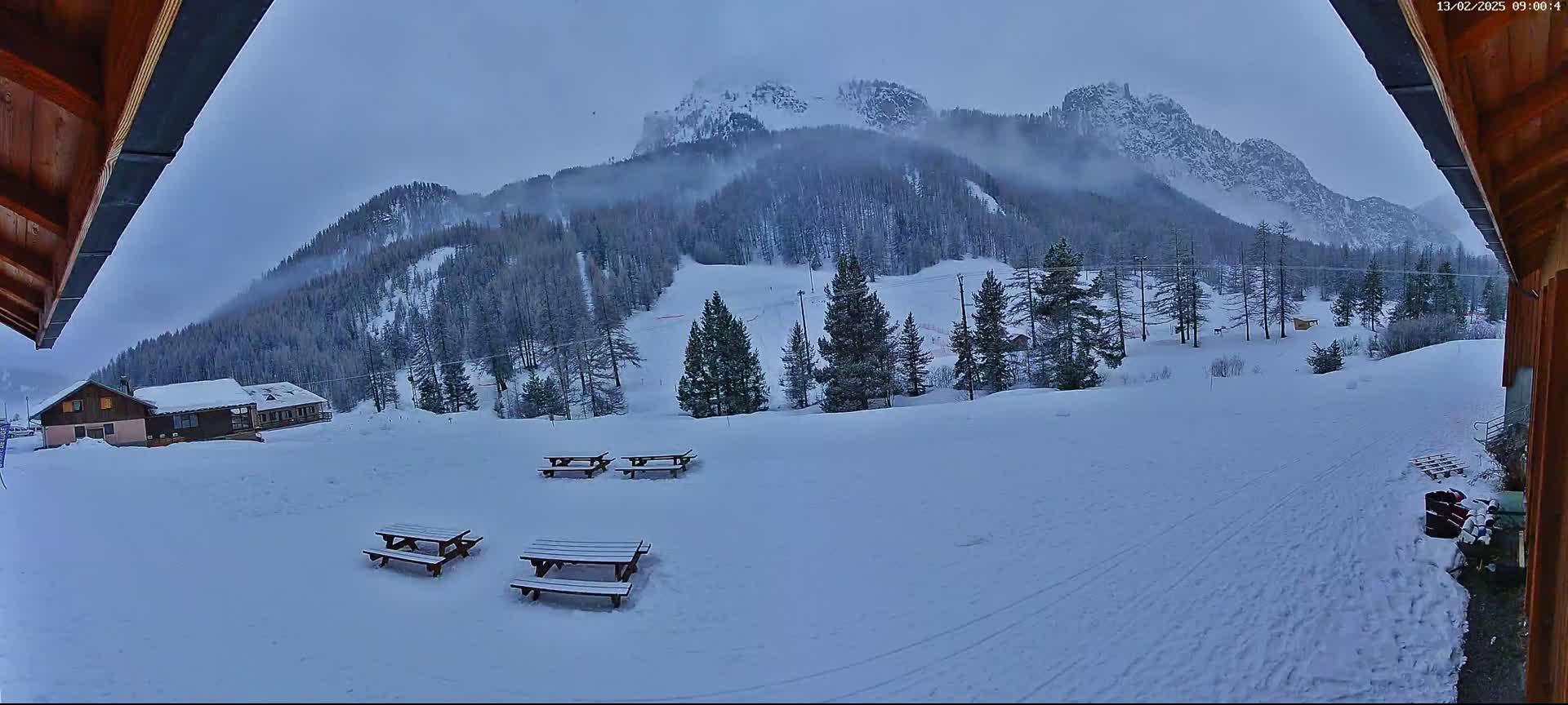 A snow-covered mountainside with several picnic tables is visible from a wooden structure's overhang under an overcast sky.