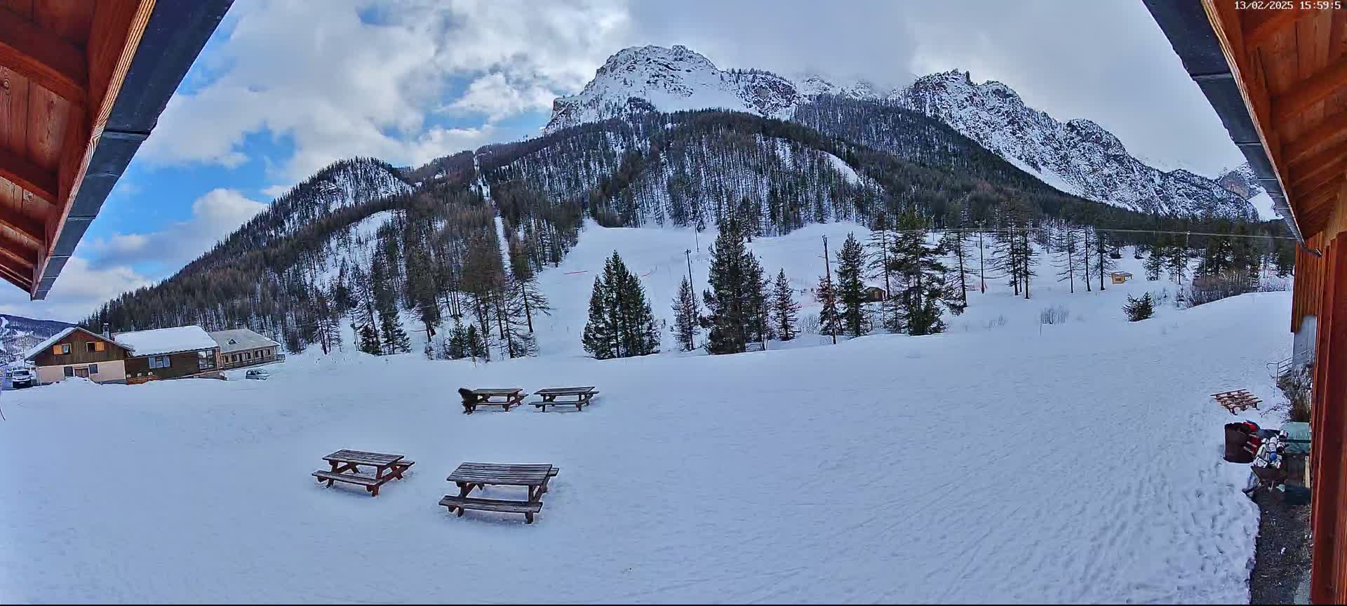 A snow-covered landscape with several picnic tables, a dog, and buildings in the foreground, overlooking a mountain range under a partly cloudy sky.