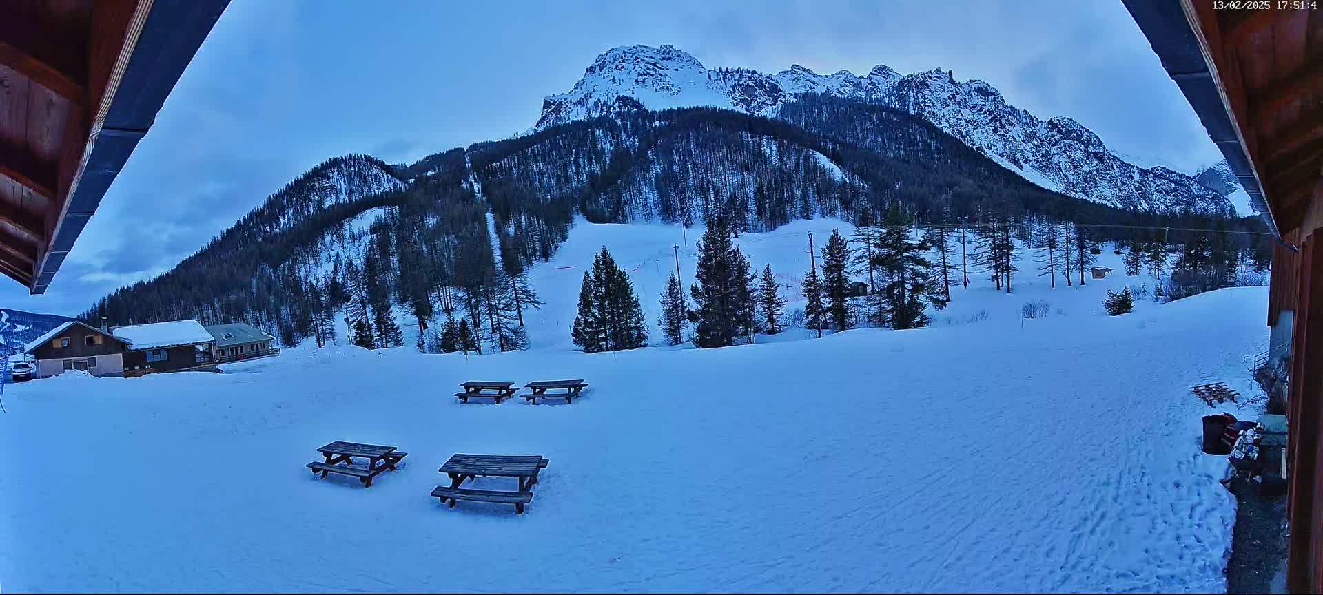 A snow-covered landscape with several picnic tables is visible in the foreground, while a mountainous area with evergreen trees is visible in the background under a twilight sky.