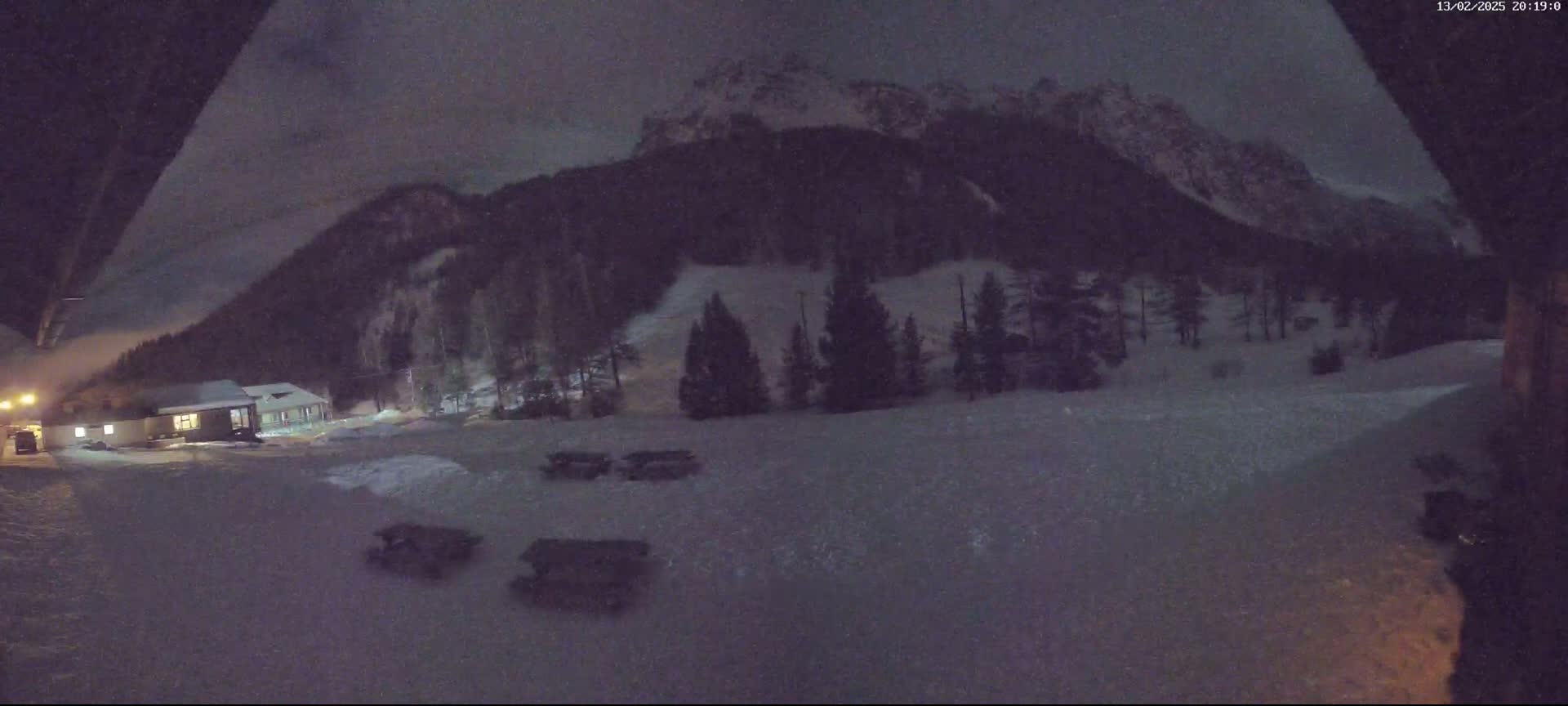A snow-covered landscape at night, featuring a small building, a stand of evergreen trees, and a snow-capped mountain in the background.