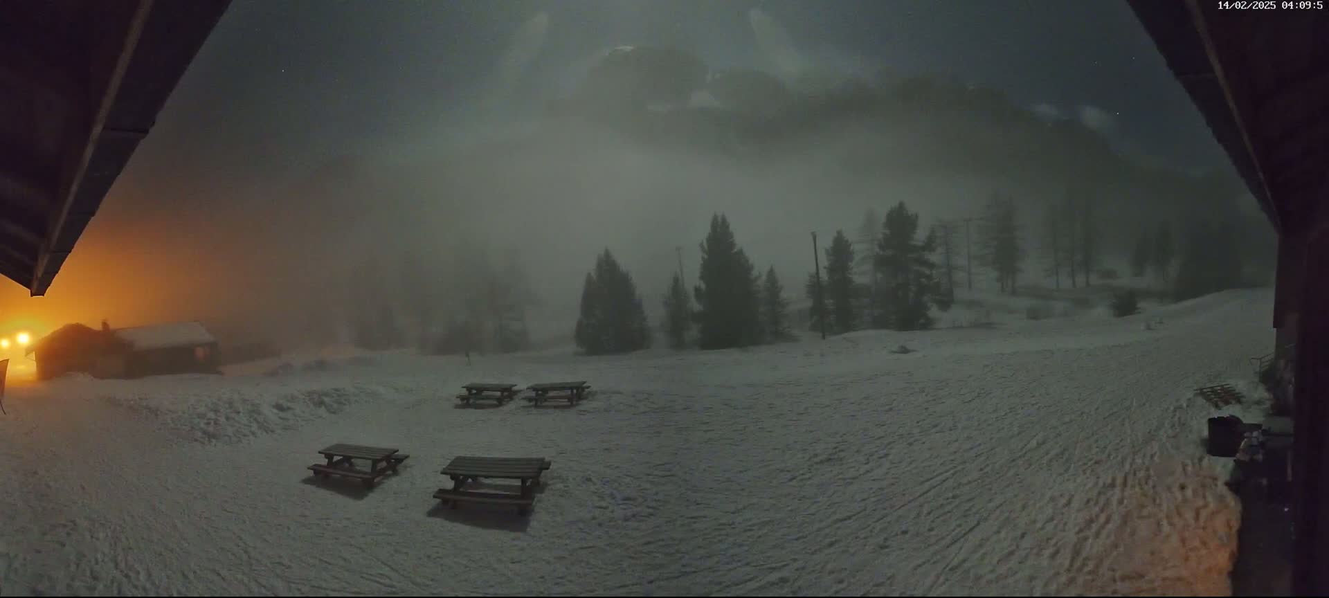 A snow-covered landscape at night is shrouded in heavy fog, with several picnic tables visible in the foreground and a small building illuminated by an orange light in the distance.