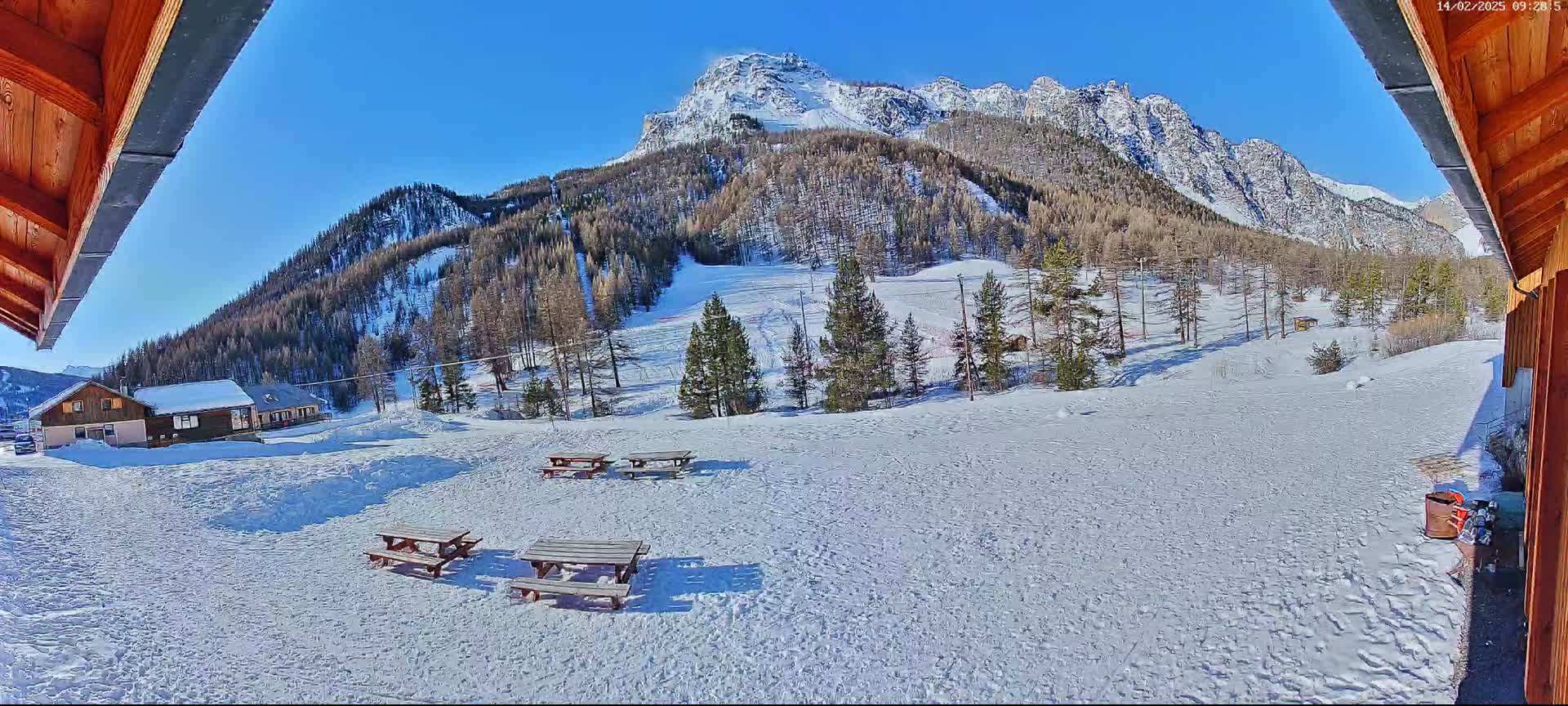 A sunny, snow-covered landscape with a few picnic tables and small buildings in the foreground, and a mountain range covered in snow and pine trees in the background.