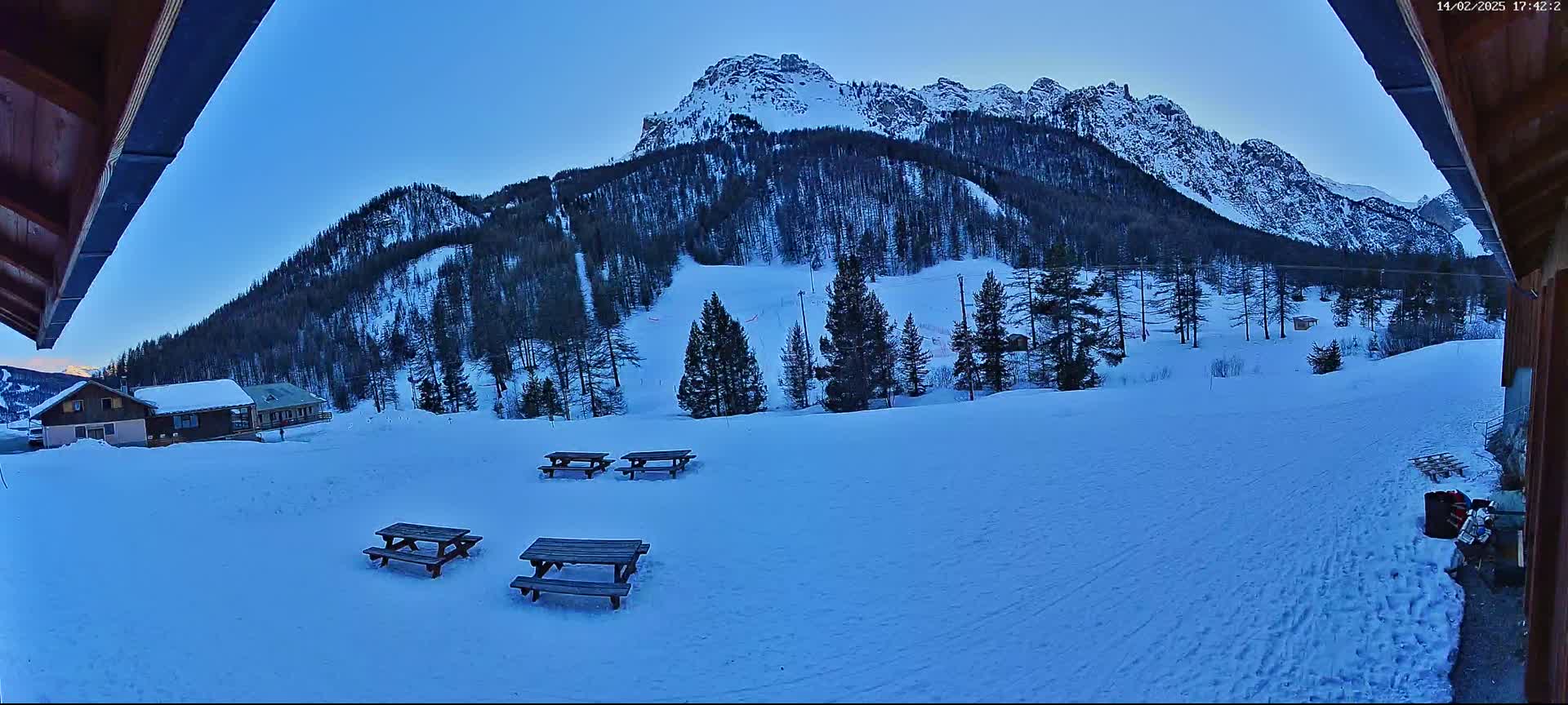 A snow-covered landscape with several picnic tables, a line of evergreen trees, and a snow-capped mountain range under a clear twilight sky.