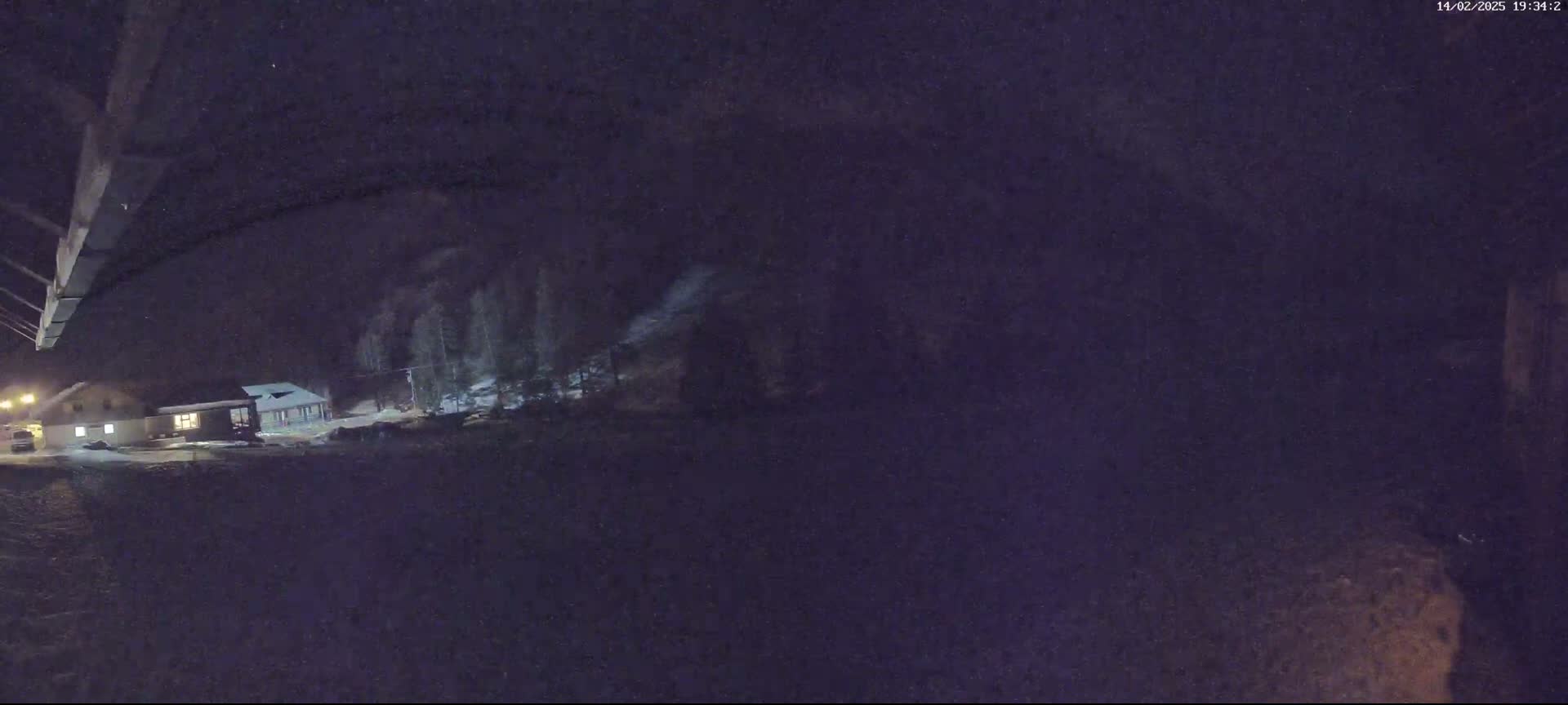A nighttime, snowy scene shows a building with lights on, a few trees, and a vehicle, viewed from under a dark structure.