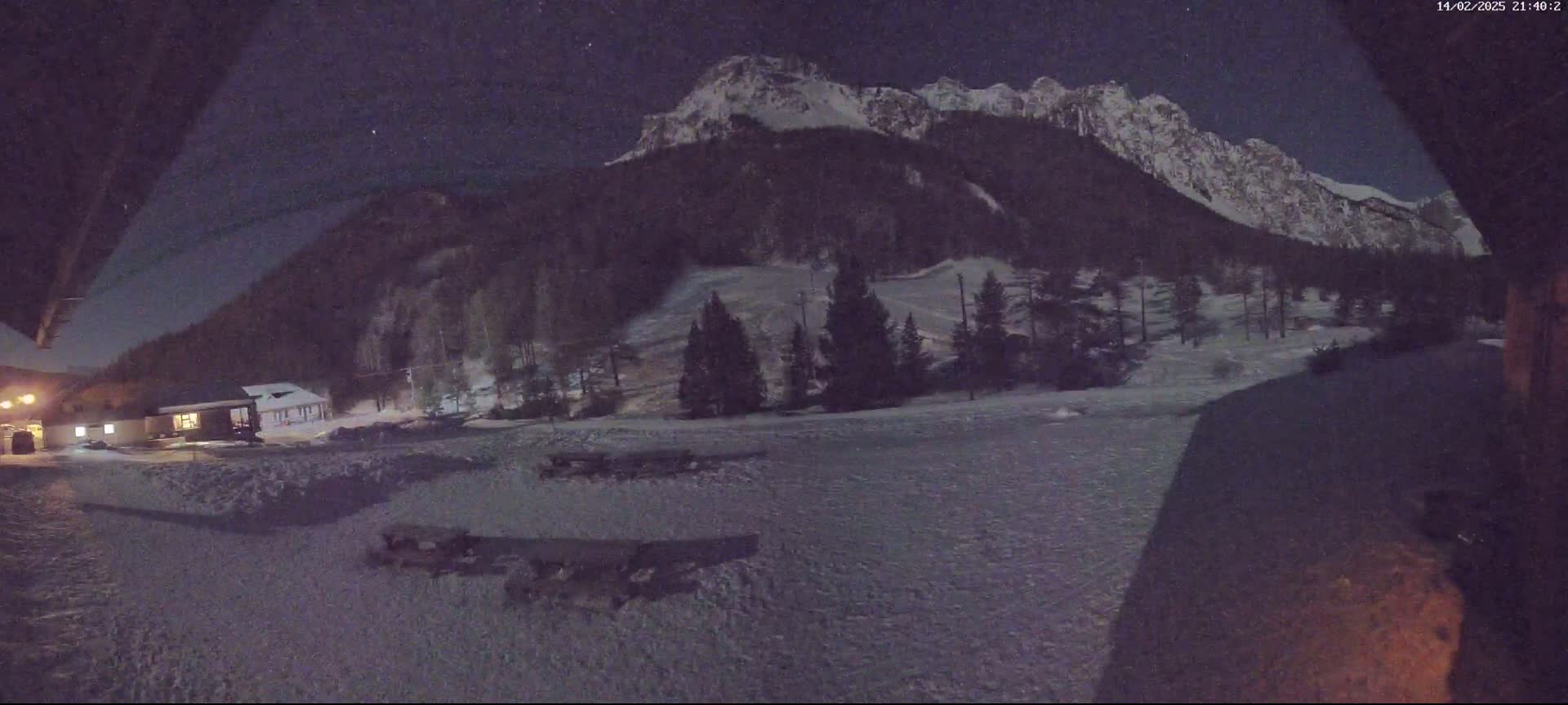 A snow-covered landscape at night is viewed from a building, showing a snow-capped mountain in the background under a starry sky.