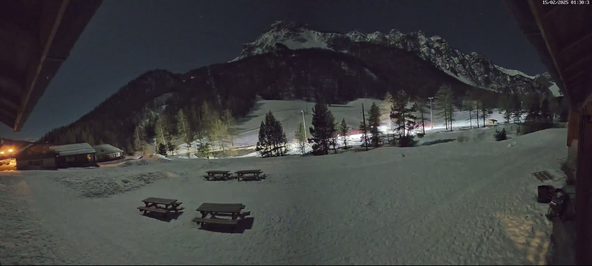 A snow-covered landscape at night, with picnic tables in the foreground and a mountain range lit by moonlight in the background.